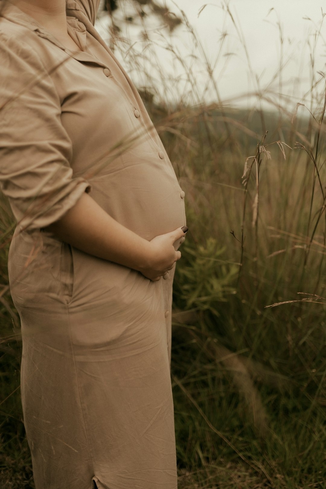 Pregnant person holding their baby bump while standing in tall grass.
